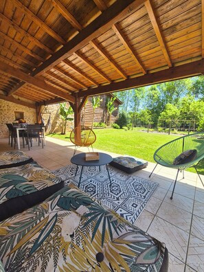 Une terrasse extérieure dotée d’un plafond en bois, d’une table à manger avec des chaises, et d’un espace gazonné agrémenté d’arbres.