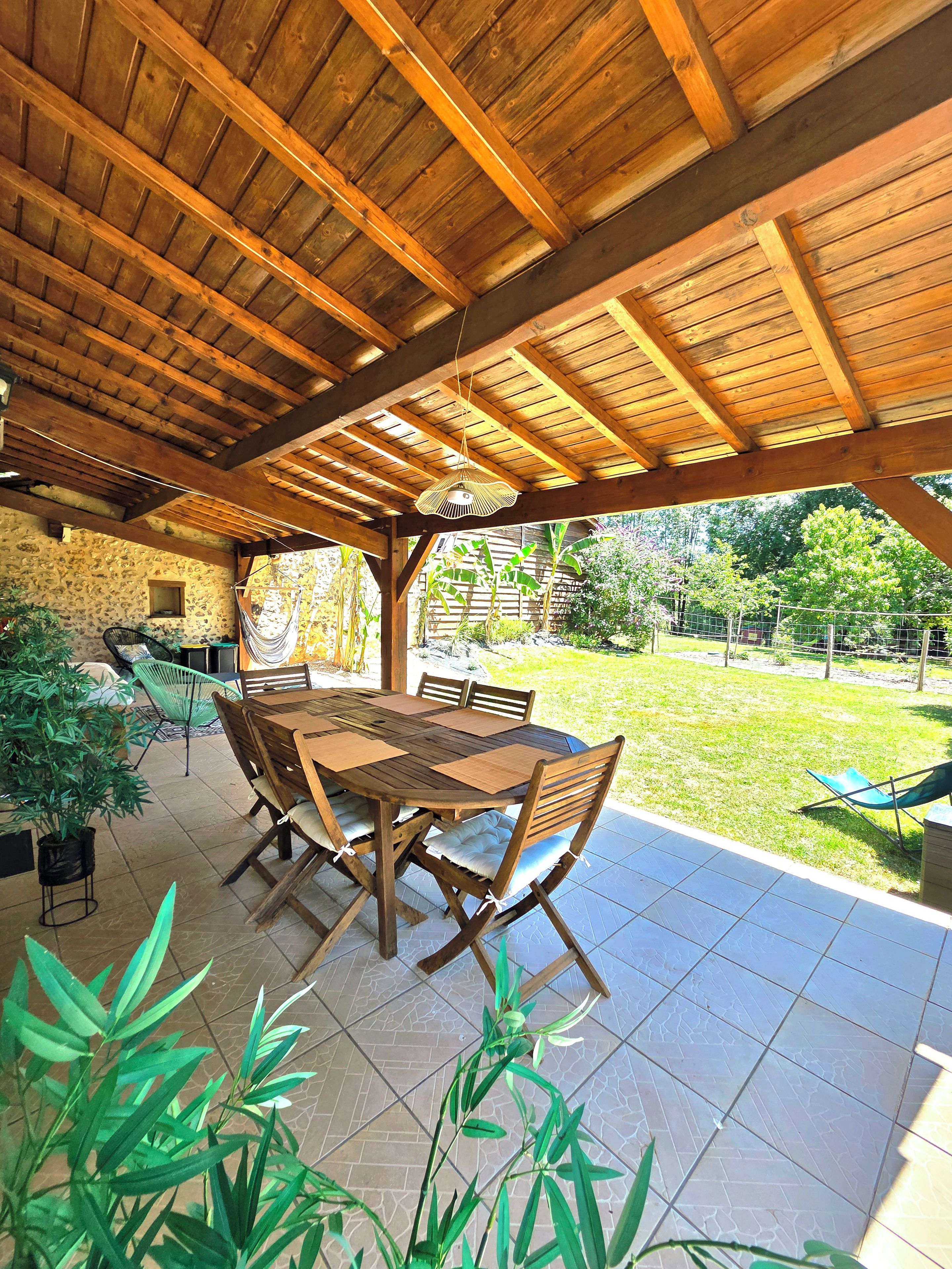 Een terras buiten met een houten tafel en stoelen, omgeven door potplanten en uitzicht op de tuin.