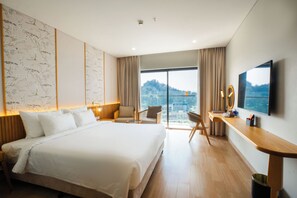 A modern hotel room with a large bed, a desk with a mirror, and a view of a cityscape through large windows. - Seabird Hotel Thien Cam (Cam Xuyen)