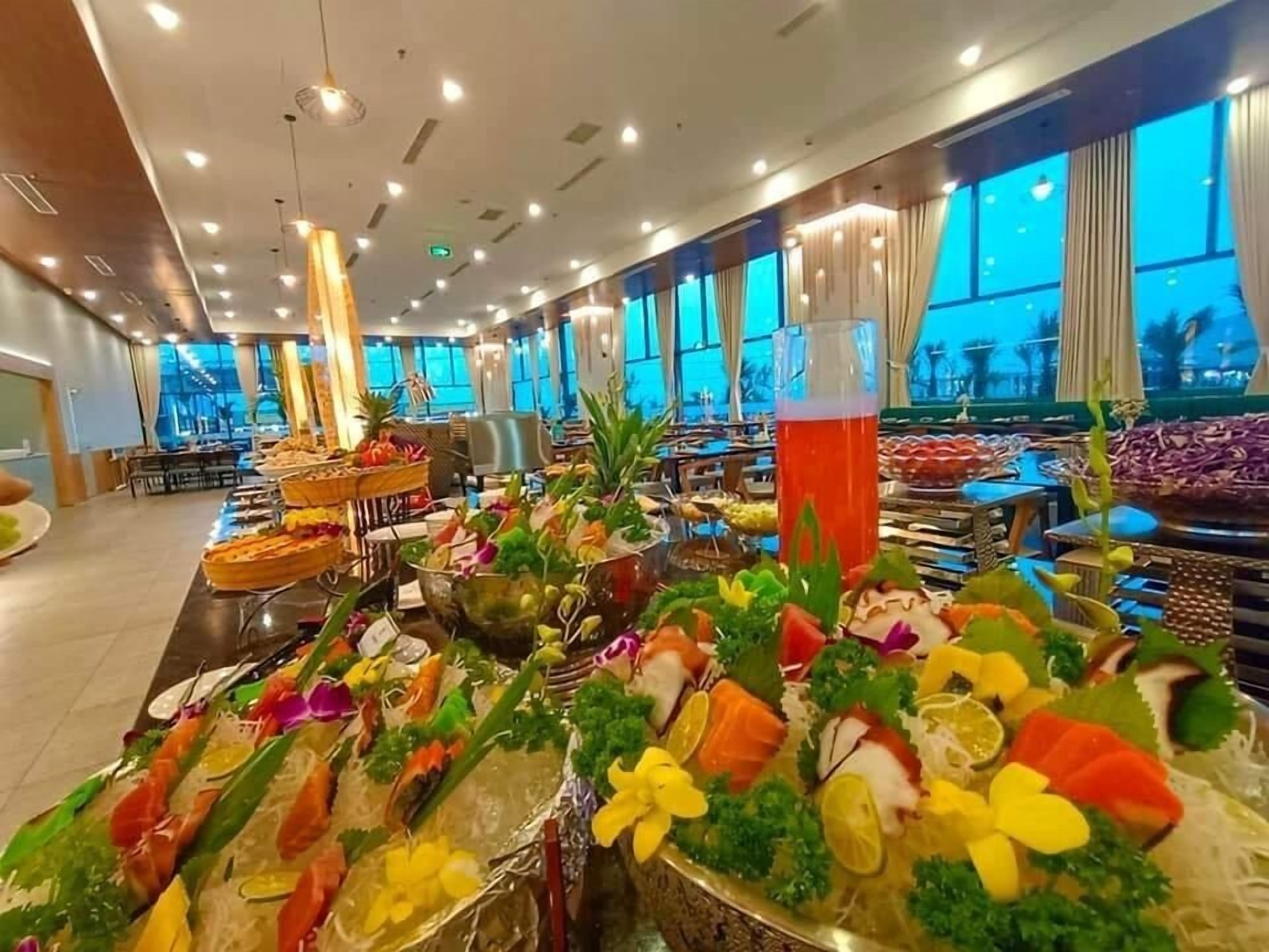 Buffet com diversos frutos do mar e frutas, decoração moderna com grandes janelas e teto com iluminação embutida.