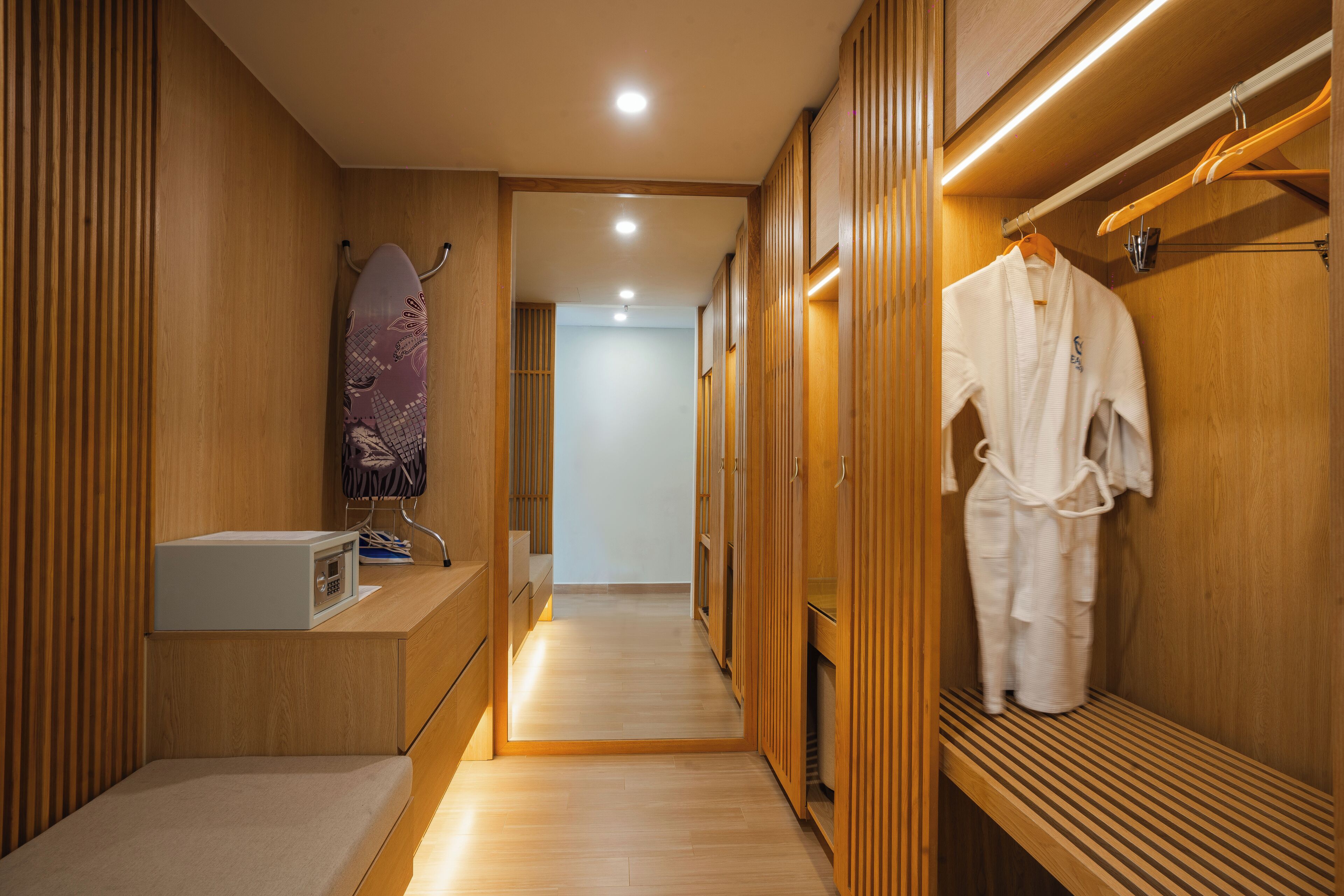 A wooden walk-in closet with a bench, a white robe hanging on a rack, and a surfboard leaning against the wall.