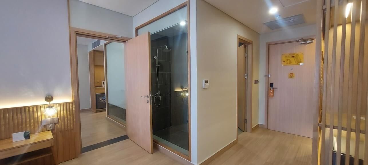 Habitación de hotel con piso de madera, cabina de ducha de vidrio, puerta de madera con un letrero y un banco con lámpara.