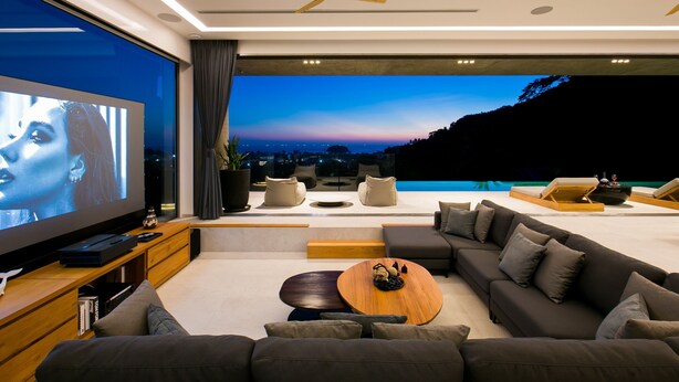 A modern living room with a large flat-screen TV, comfortable seating, a wooden coffee table, and a view of a sunset over a body of water. - Villa Nordic Star Seaview Near Kata Beach (Muang)