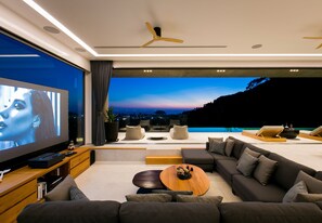 A modern living room with a large flat-screen TV, comfortable seating, a wooden coffee table, and a view of a sunset over a body of water. - Villa Nordic Star Seaview Near Kata Beach (Muang)