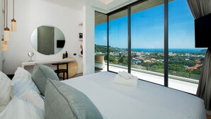 A modern hotel room with a large bed, a round mirror, a desk, and a view of a cityscape through large windows. - Villa Nordic Star Seaview Near Kata Beach (Muang)