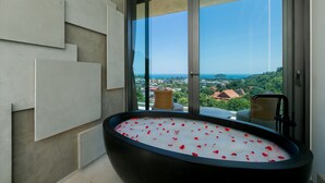 A modern bathroom with a large bathtub filled with water and rose petals, a view of a cityscape through large windows, and a balcony with a wicker chair. - Villa Nordic Star Seaview Near Kata Beach (Muang)