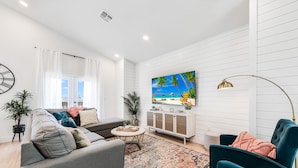 A modern living room with a sofa, armchairs, a coffee table, a TV, and a painting of a beach scene.