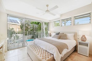 Un dormitorio con una cama grande, un banco, un ventilador de techo y vistas a una piscina y follaje exterior.