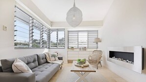 Een moderne woonkamer met een grijze bank, een bijzettafeltje, een rieten fauteuil, een open haard en grote ramen met zonwering.