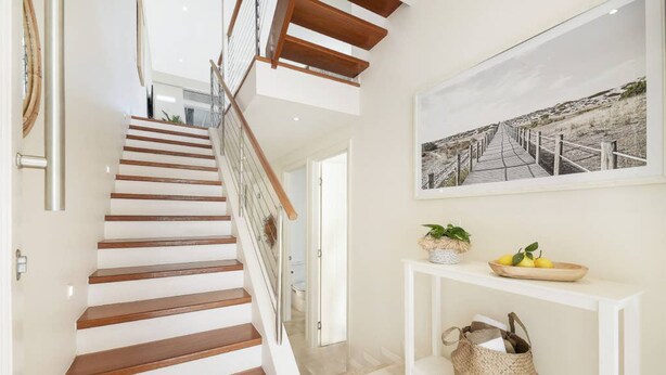 Een trap met houten treden en een glazen balustrade, die leidt naar een hal met een witte console tafel en een ingelijste foto aan de muur.