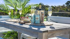 Een houten tafel met een fles champagne, twee glazen, een fles wijn en een schaal fruit op een balkon met uitzicht op zee.