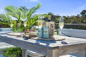 Una mesa de madera con una botella de champán, dos copas, una botella de vino y un plato de fruta en un balcón con vistas al mar.