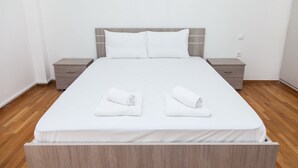 A neatly made bed with white linens and two rolled towels on the bed. There are two bedside tables with drawers, one on each side of the bed. The room has wooden flooring and white walls. - Glamorous Apt in central Athens (Athens)