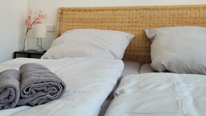 A neatly made bed with white linens and a rolled gray towel, a wicker headboard, a bedside table with a lamp and a vase of flowers.