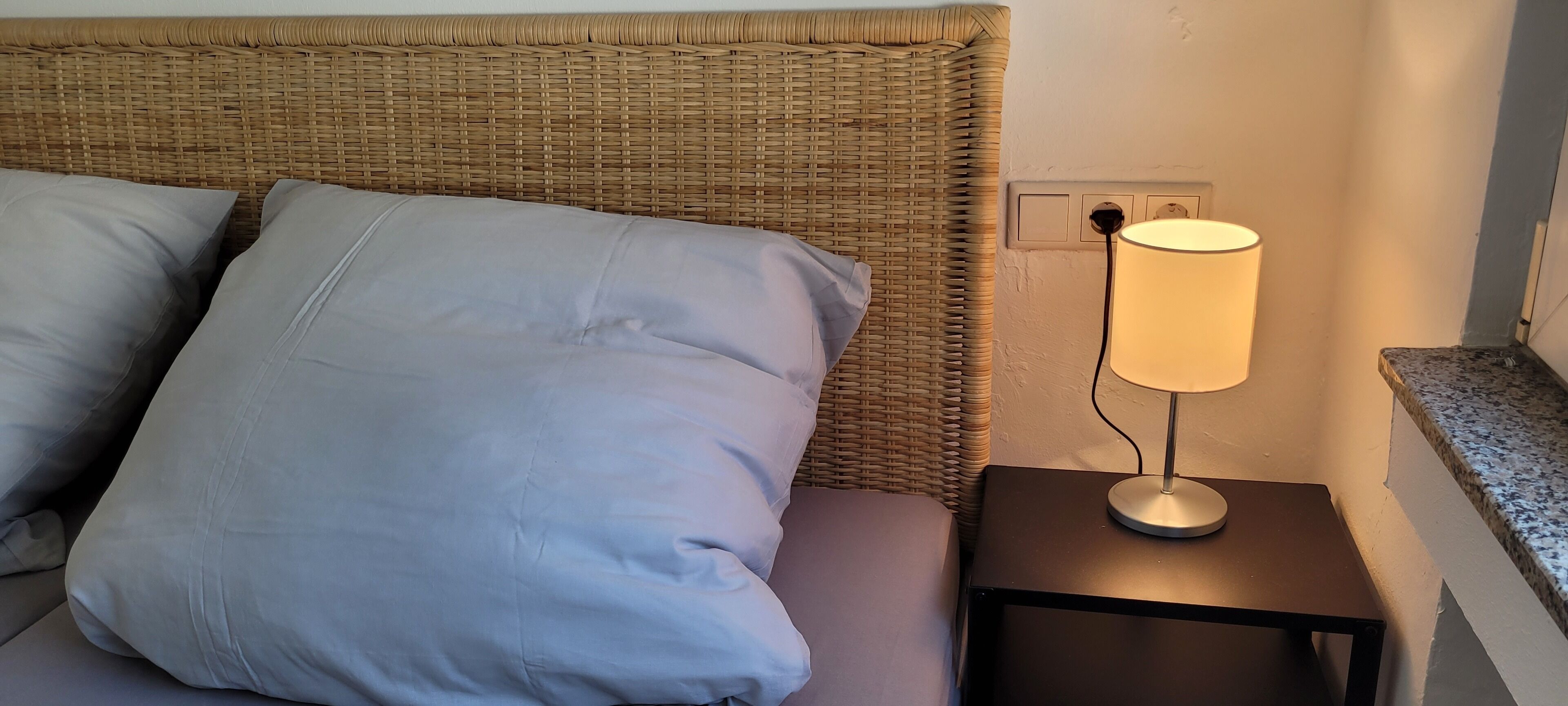 Una cama con almohadas grises y cabecera de mimbre, una mesita de noche con lámpara y encimera de granito.