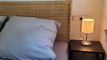 Una cama con almohadas grises y cabecera de mimbre, una mesita de noche con lámpara y encimera de granito.