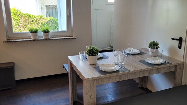 A dining area with a wooden table set for four, potted plants on the windowsill, and a welcome sign on the wall.