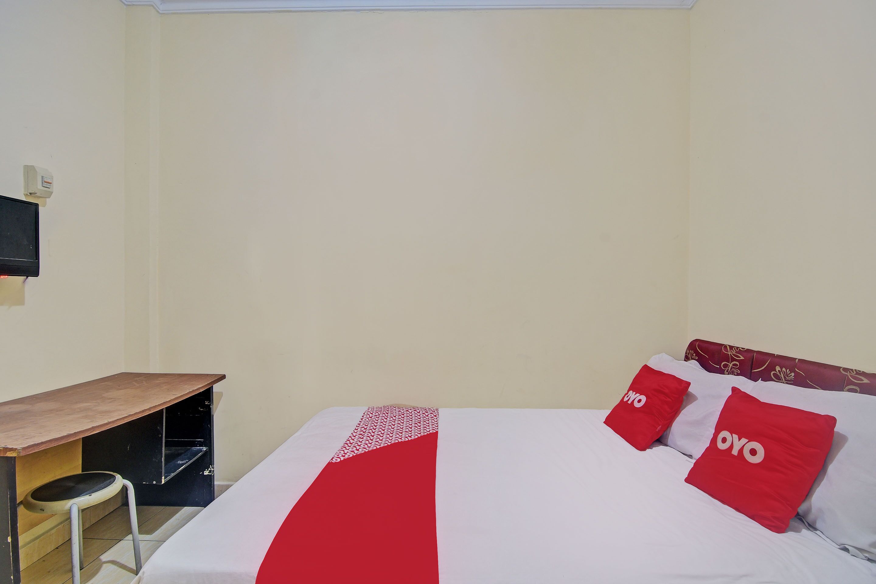 A hotel room with a bed, two red pillows with the word "OYO" on them, a wooden desk, and a small stool.