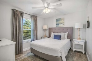 Quarto com cama, duas mesas de cabeceira, ventilador no teto, janela com cortinas e um quadro na parede.