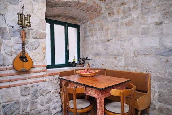 Una habitación rústica con paredes de piedra, una mesa y sillas de madera, un ventanal con contraventanas verdes y un instrumento musical de cuerda tradicional colgado en la pared.