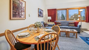 A dining area with a round wooden table set for four, surrounded by wooden chairs. There is a sofa with patterned cushions in the background, a floor lamp, and a window with curtains. - Captivating Condo with sports & fitness center, pool, and hot tub - Highridge E10 (Killington)