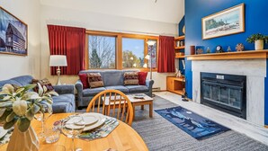 A living room with a fireplace, a sofa, a dining table set, and a large window with curtains. - Captivating Condo with sports & fitness center, pool, and hot tub - Highridge E10 (Killington)