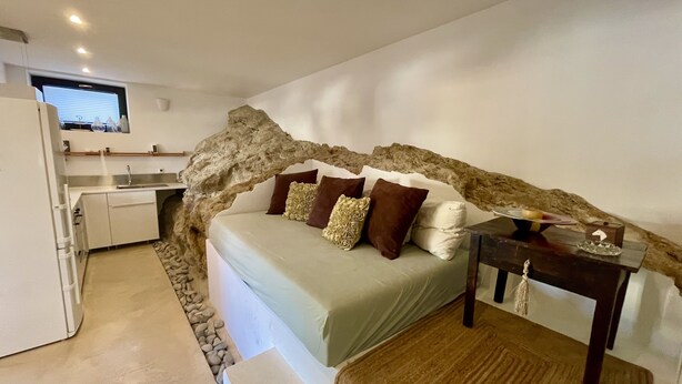 A bedroom with a stone arch, a bed with rose-patterned pillows, a small table with a decorative item, and a kitchenette in the background.