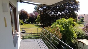 A balcony with a metal railing, a covered area, and a view of a garden and trees.