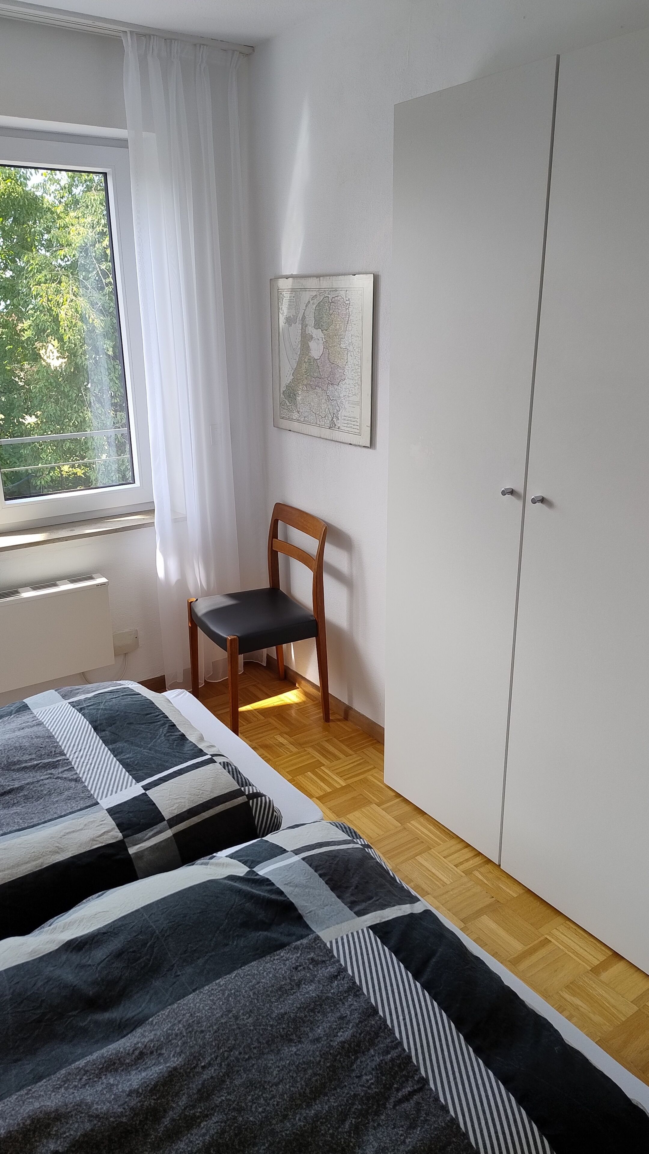 A bedroom with two beds, a chair, a wardrobe, a map on the wall, and a window with curtains.
