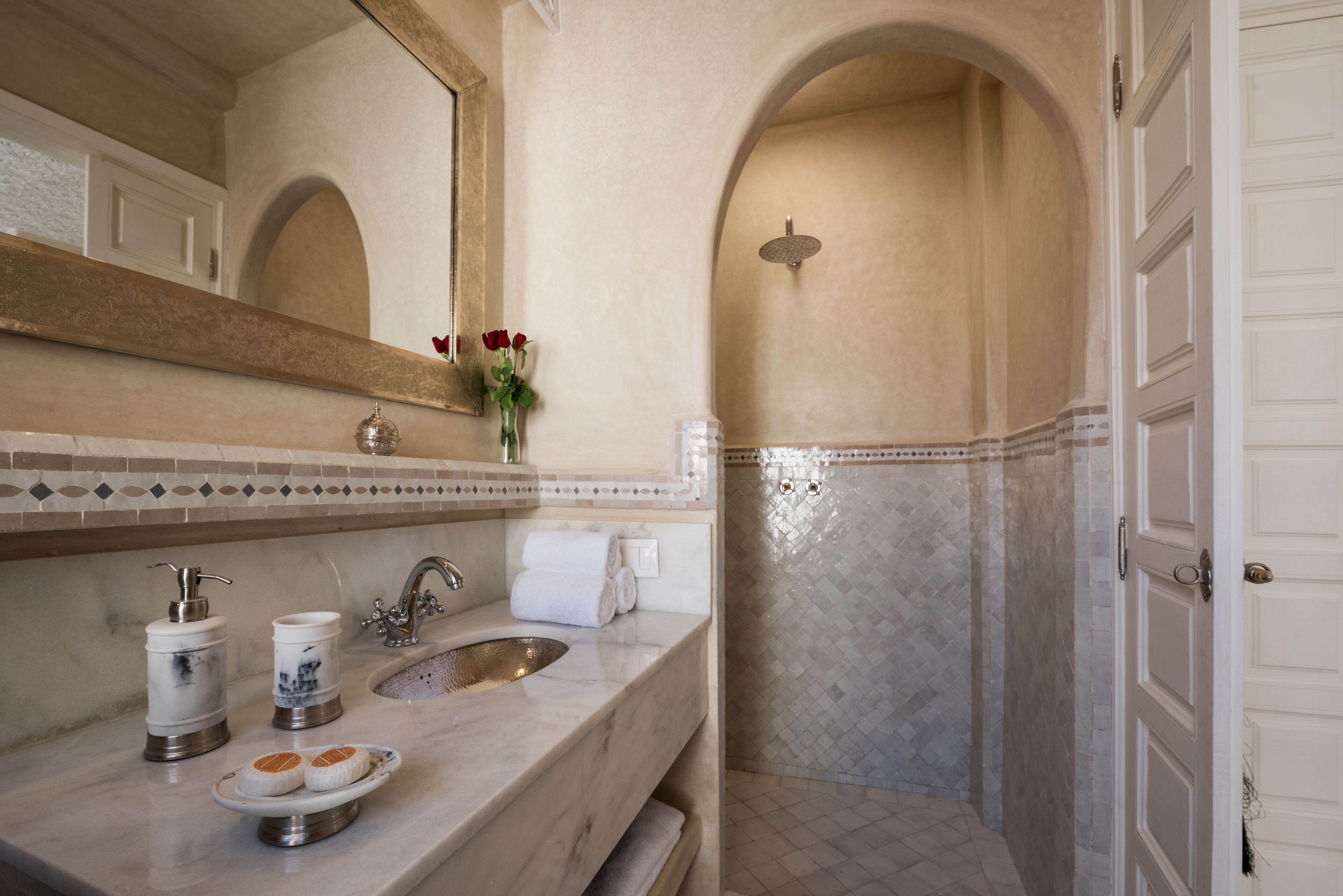 Een badkamer met een marmeren wastafel, een spiegel, een douche en een betegelde muur.