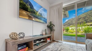 Ein Wohnzimmer mit einem großen Fernseher, einem Holztisch mit Büchern, einer Pflanze und Blick auf eine Berglandschaft durch eine Schiebetür.