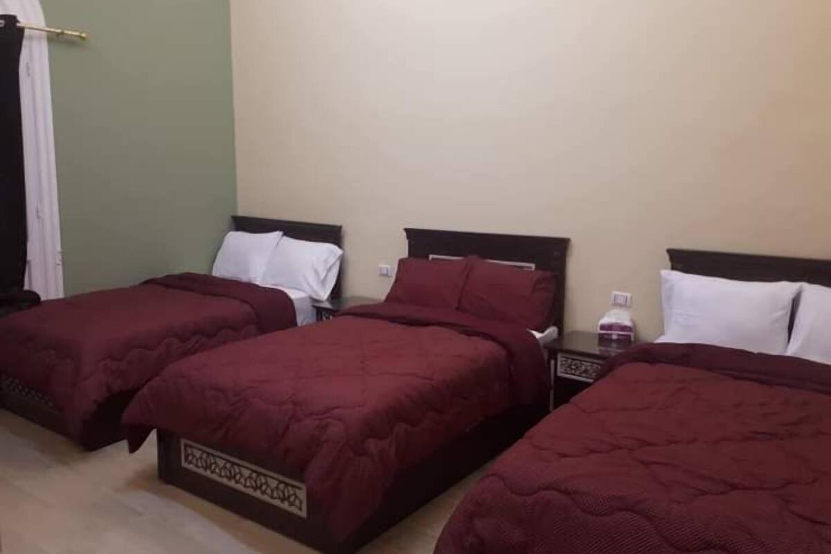 A hotel room with three single beds, each with a dark headboard and white pillows. The room has a green wall and a wooden floor.