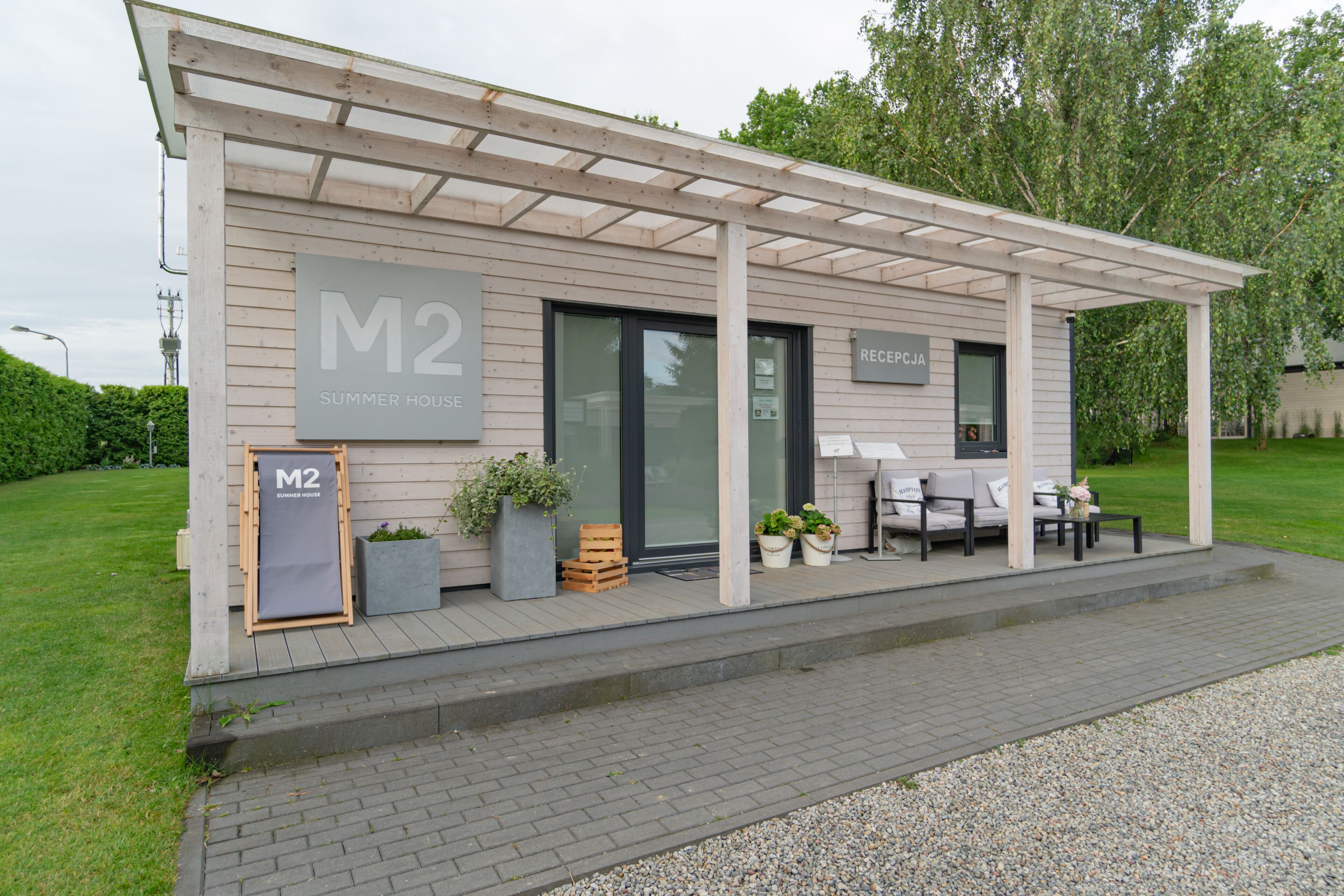 A modern building with a covered patio area, labeled "M2 Summer House," featuring outdoor seating and potted plants.