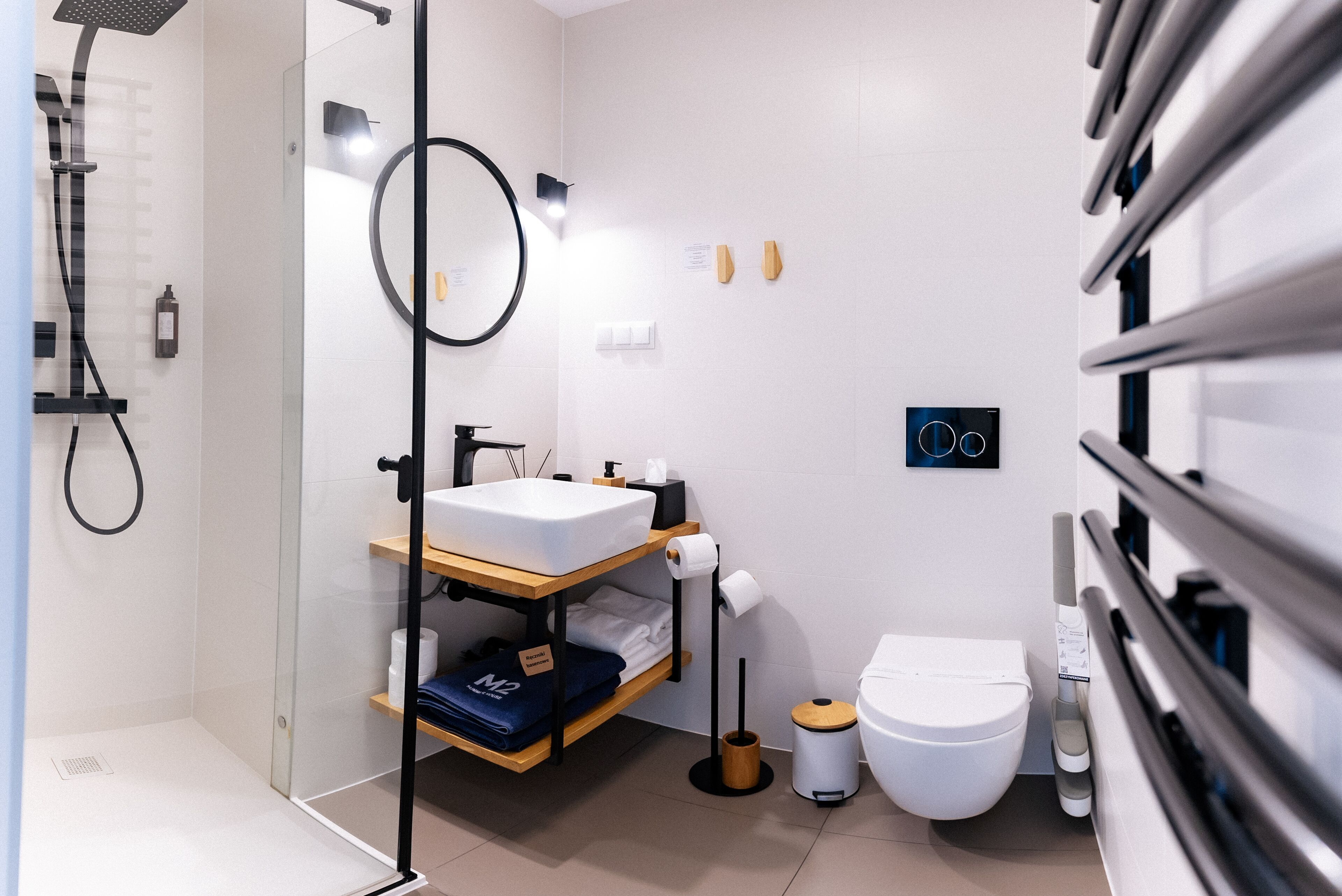 Une salle de bain moderne avec une douche à parois de verre, un lavabo doté d’une étagère en bois, des toilettes et une poubelle.