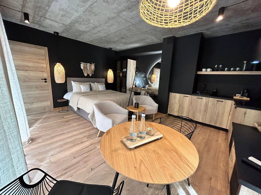 Um quarto moderno com uma cama grande, mesas de cabeceira, uma escrivaninha, uma mesa de jantar redonda com copos e garrafas, e uma cozinha compacta.