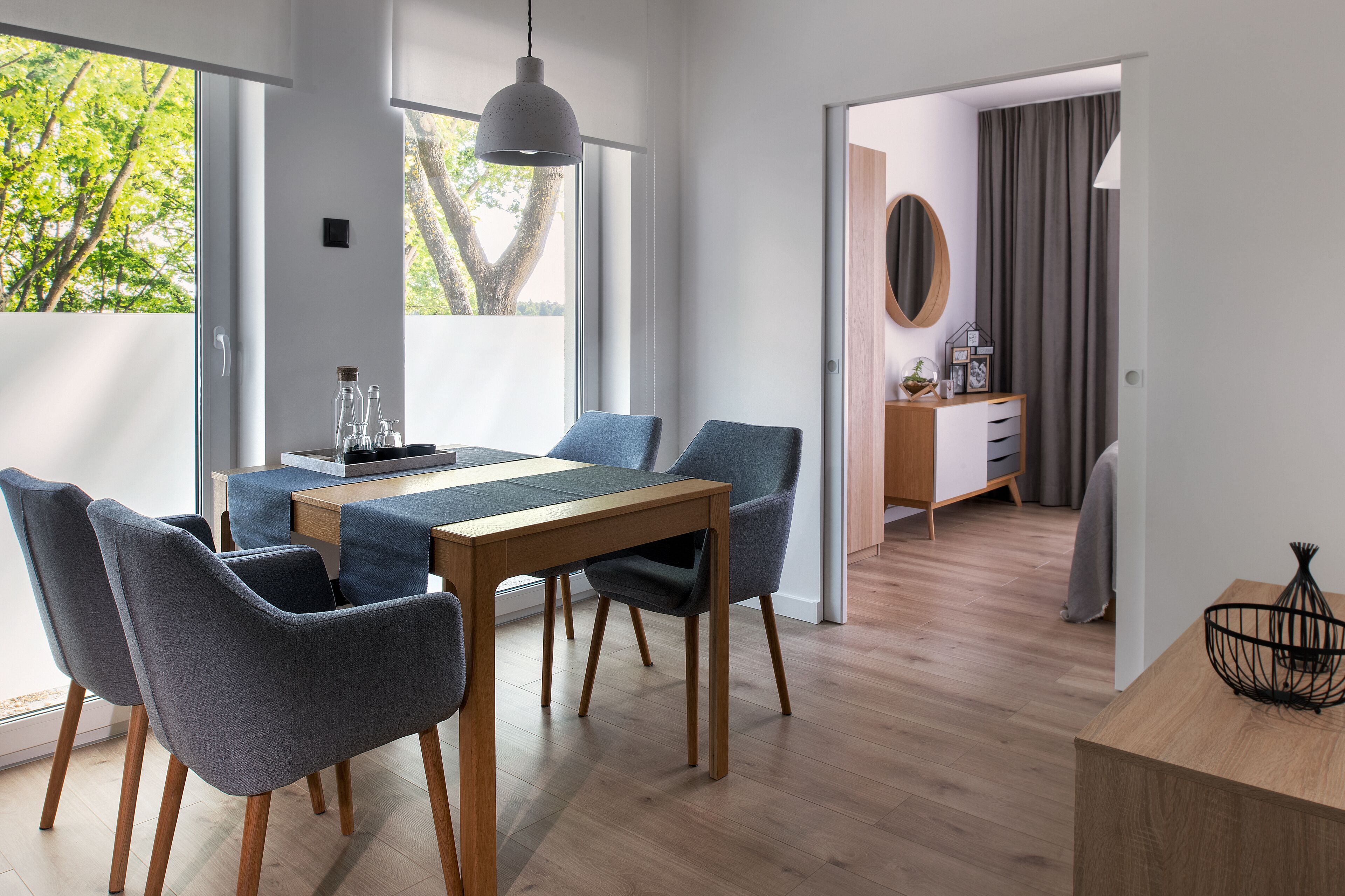 Un comedor moderno con una mesa de madera y sillas grises tapizadas, una lámpara colgante y vistas al exterior.