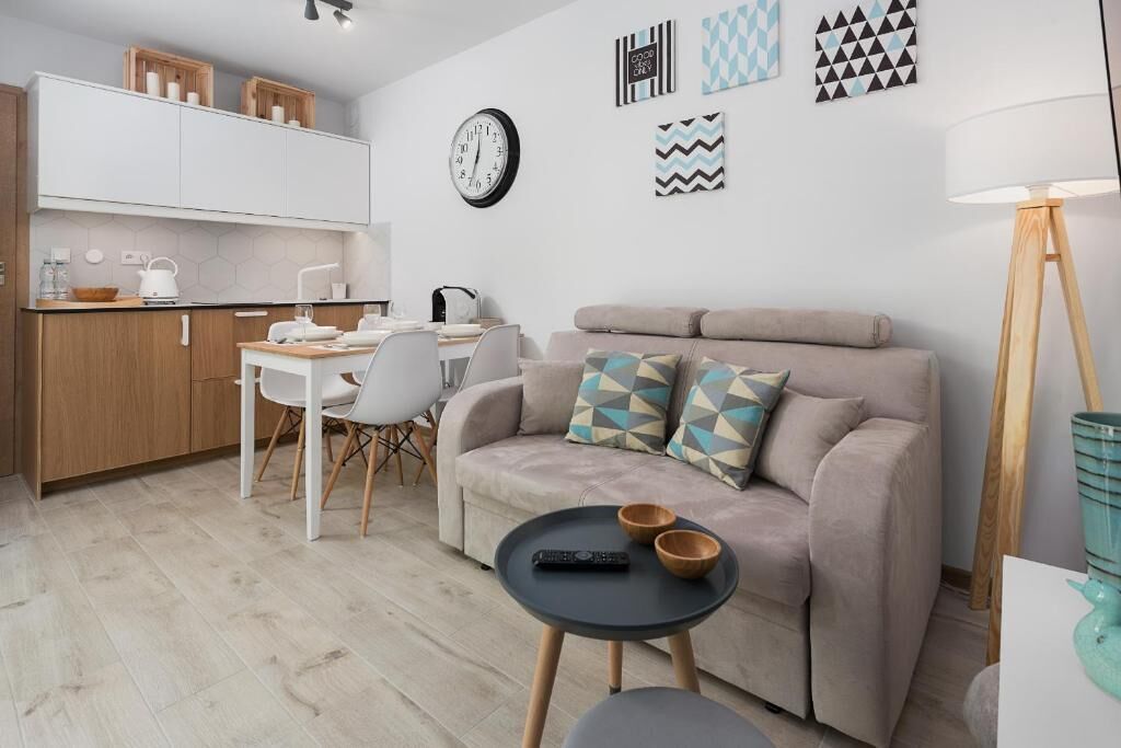 Un espacio habitable compacto con kitchenette, un sofá con cojines estampados, una zona de comedor con mesa y sillas, y una mesita auxiliar con cuencos y un control remoto.