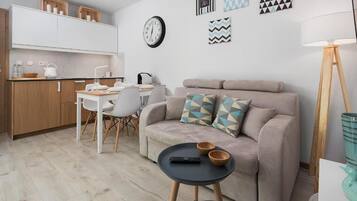 Un espacio habitable compacto con kitchenette, un sofá con cojines estampados, una zona de comedor con mesa y sillas, y una mesita auxiliar con cuencos y un control remoto.