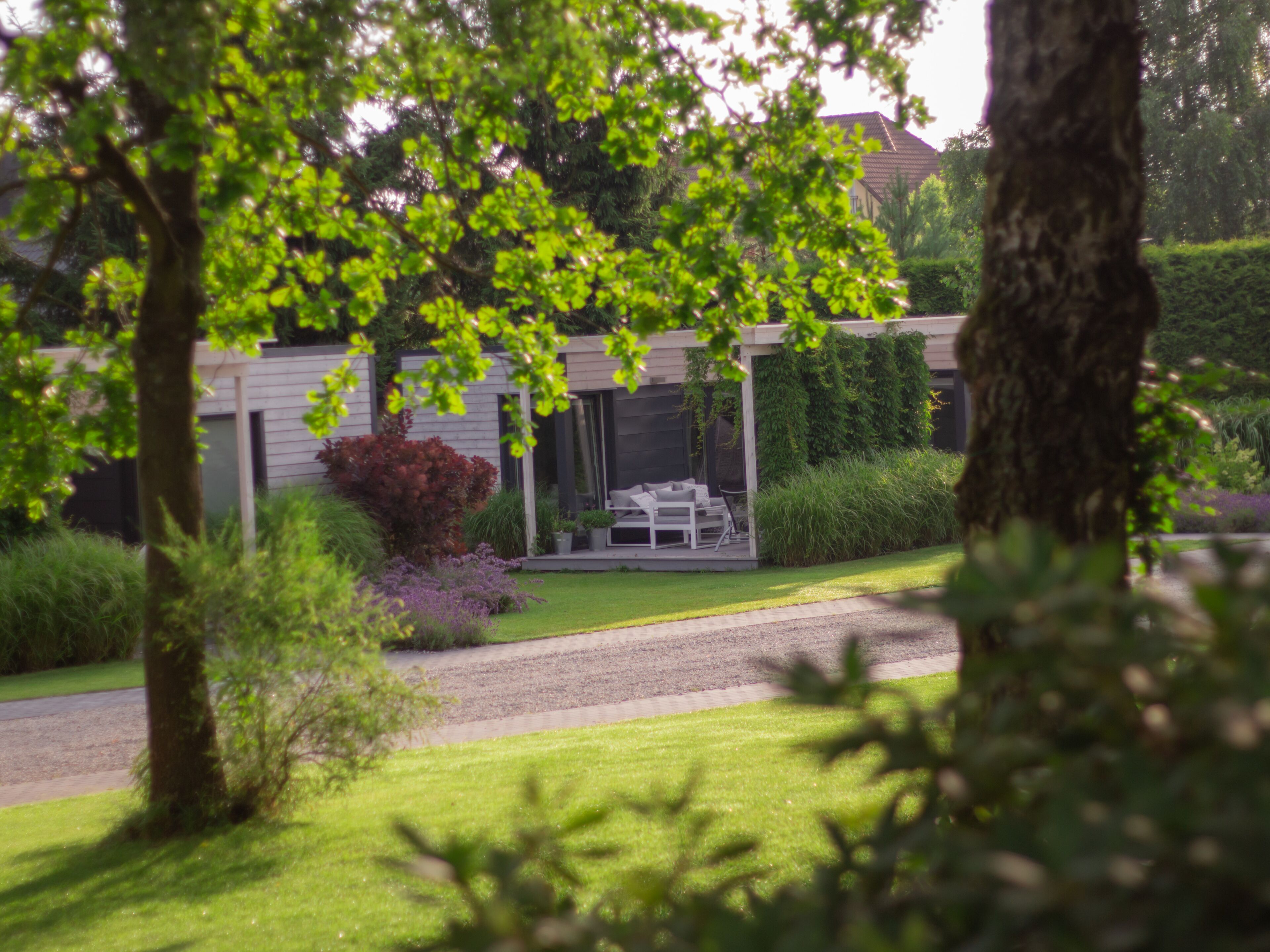 Et moderne hus med et overdækket udendørs opholdsområde, omgivet af frodig vegetation og en velplejet græsplæne.