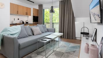 Una sala de estar moderna con un sofá gris, una mesita y un televisor. Se observa una cocina al fondo con armarios de madera y un ventanal.