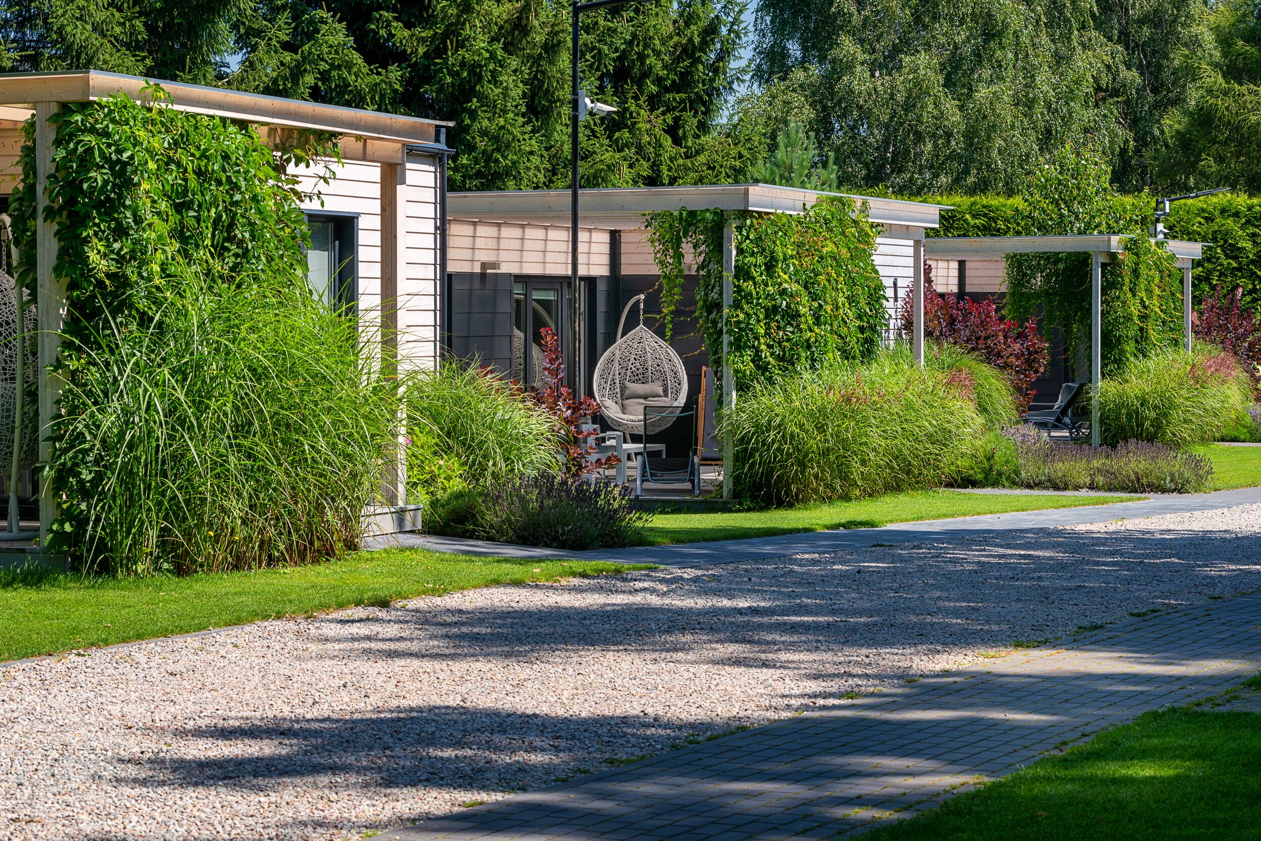 Ein modernes Gebäude mit überdachter Außenbestuhlung, umgeben von üppiger Bepflanzung und einem Kiesweg.