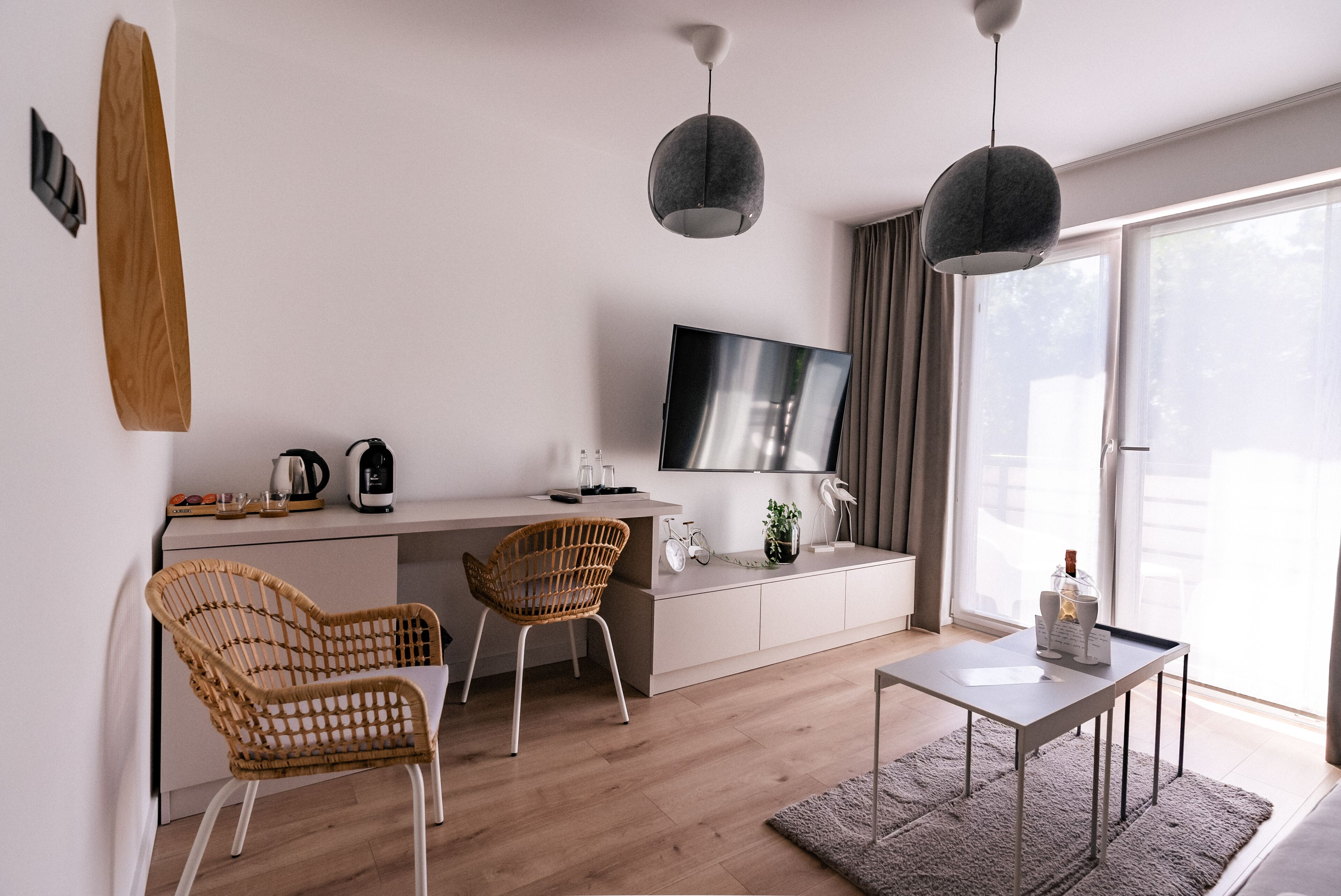 Ein modernes Wohnzimmer mit Flachbildfernseher, zwei Korbstühlen, einem kleinen Tisch und einem großen Fenster mit Vorhängen.