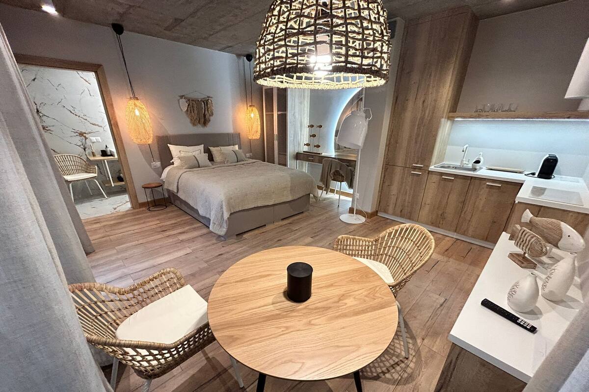 Une chambre moderne comprenant un lit, des tables de chevet, une table ronde en bois, des chaises en osier, un luminaire suspendu et un coin cuisine avec un évier et des placards.
