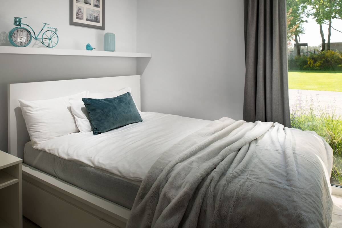 Un lit bien fait, recouvert d’une couverture grise, avec un oreiller blanc et un autre d’une couleur turquoise. Une table de chevet blanche se trouve à côté du lit. Au-dessus du lit, on aperçoit une étagère sur laquelle sont posés un vélo turquoise, une petite figurine et un tableau encadré.