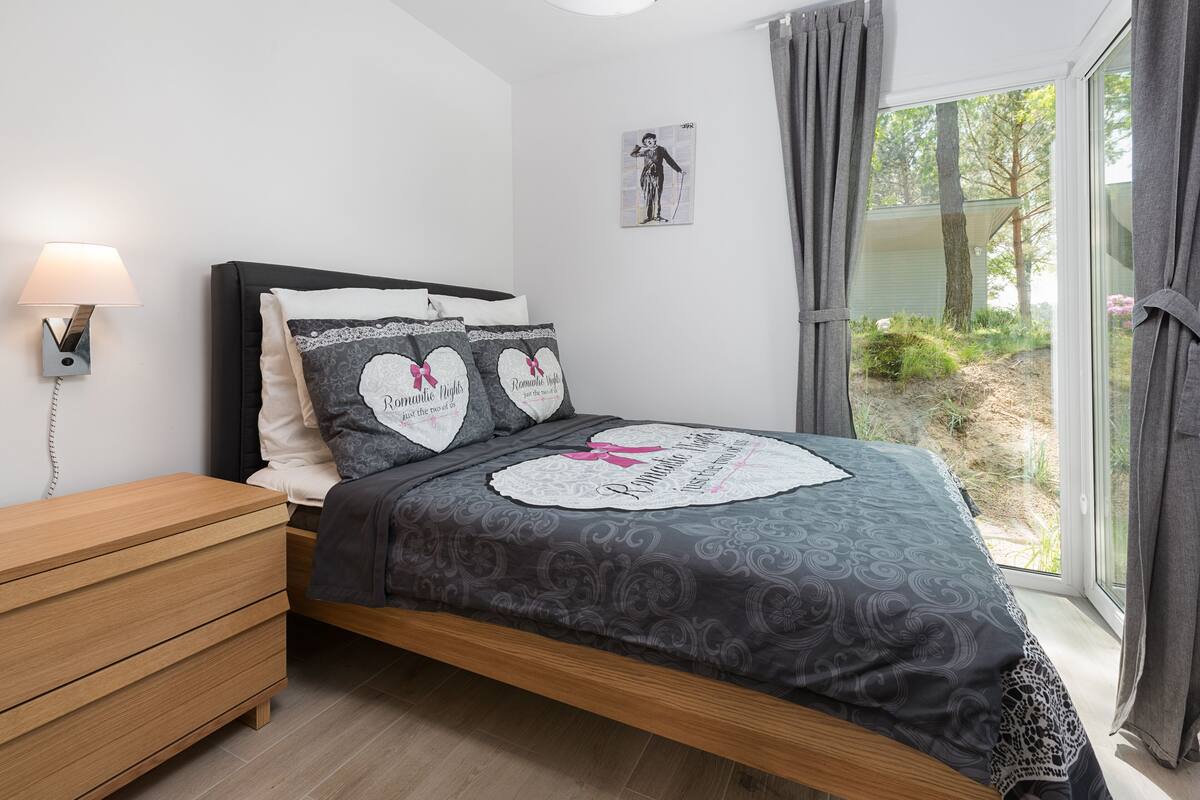 Une chambre à coucher avec un lit, une table de chevet, une lampe, un tableau encadré au mur et une vue sur la verdure à l’extérieur.