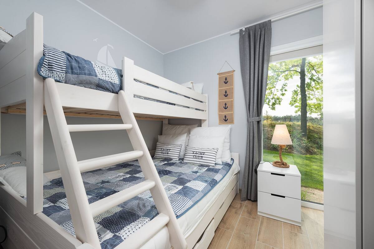 Un lit superposé avec une échelle, une table de chevet avec une lampe et une vue sur un jardin par la fenêtre.