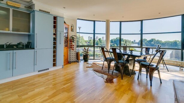 A modern kitchen with a view of a river, a dining area with a table and chairs, and a large window overlooking the scenery. - Brand New 2-bed Apartment in Brentford Kew Gardens (Brentford)