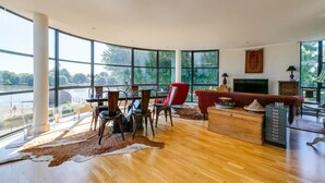 A spacious living room with a large window overlooking a river, a wooden dining table with chairs, a red armchair, and a fireplace. - Brand New 2-bed Apartment in Brentford Kew Gardens (Brentford)
