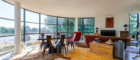 A spacious living room with a large window overlooking a river, a wooden dining table with chairs, a red armchair, and a fireplace.