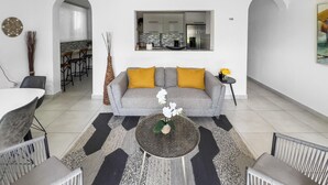 Un soggiorno moderno con un divano grigio, cuscini gialli, un tavolino rotondo con un vaso di fiori e una cucina visibile attraverso un arco.